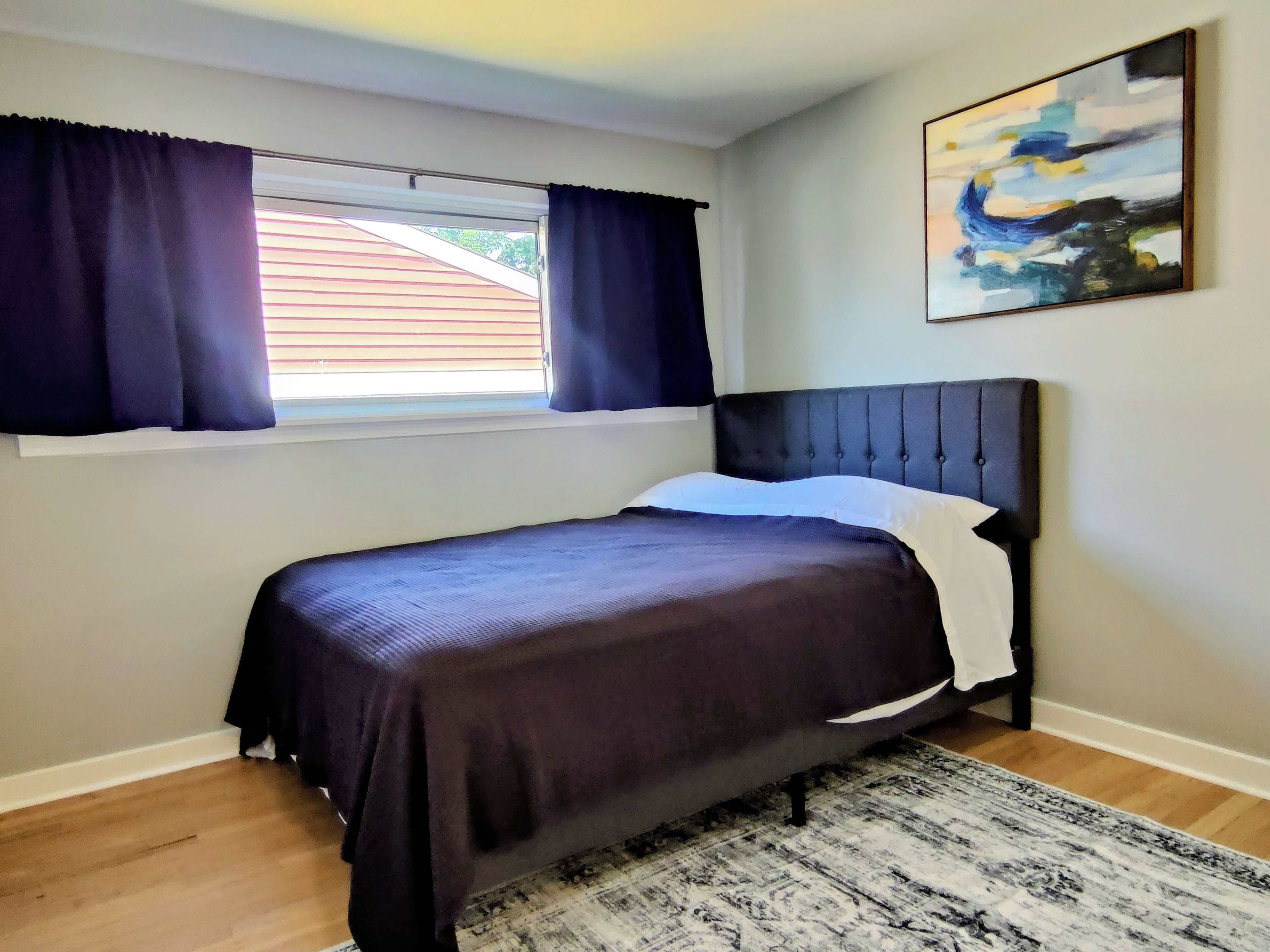 A bedroom with a bed, a window with curtains, a painting on the wall, and a patterned rug.