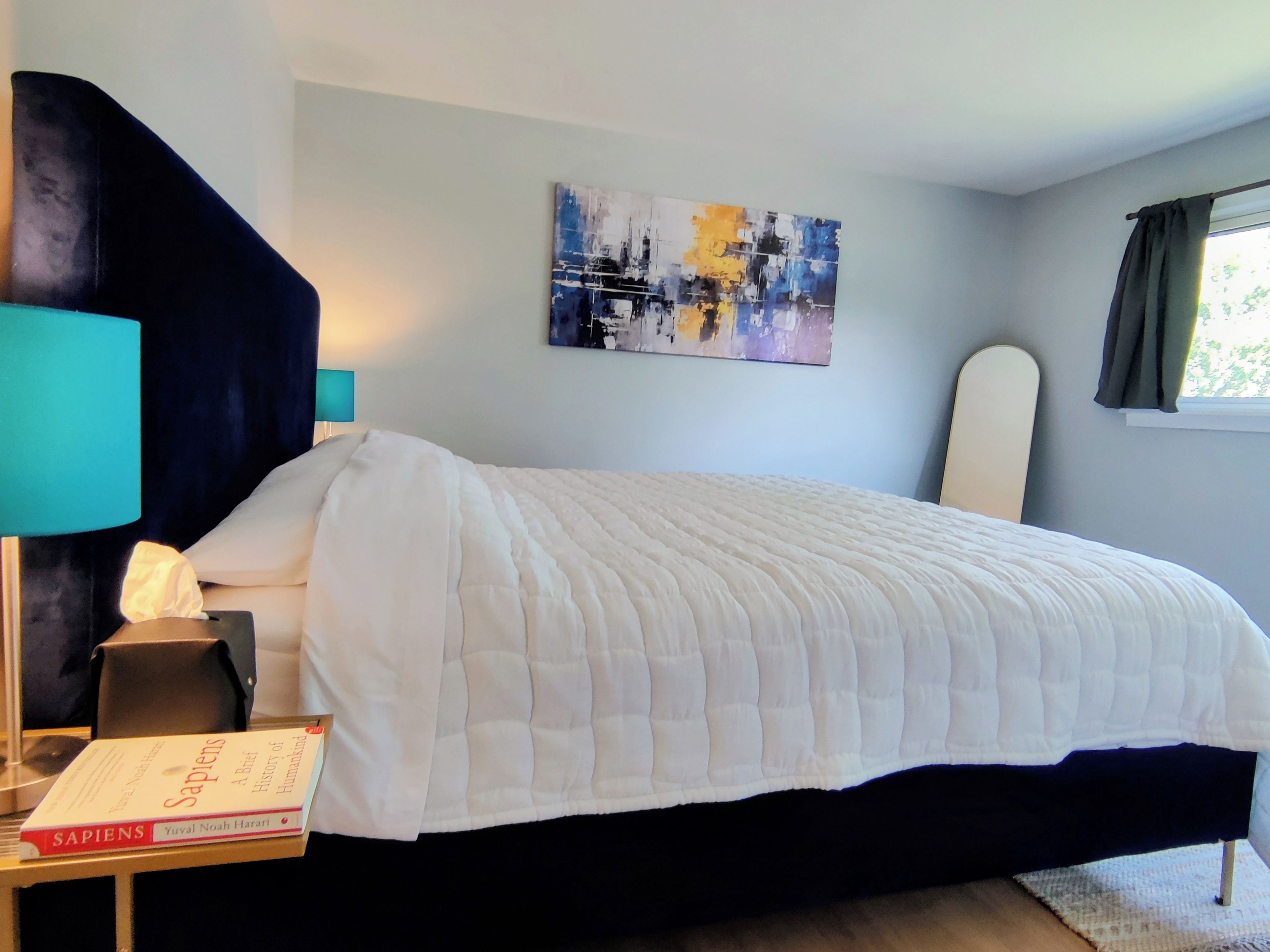A neatly made bed with a white duvet, a bedside lamp, a book titled 'Sapiens,' and a painting on the wall.