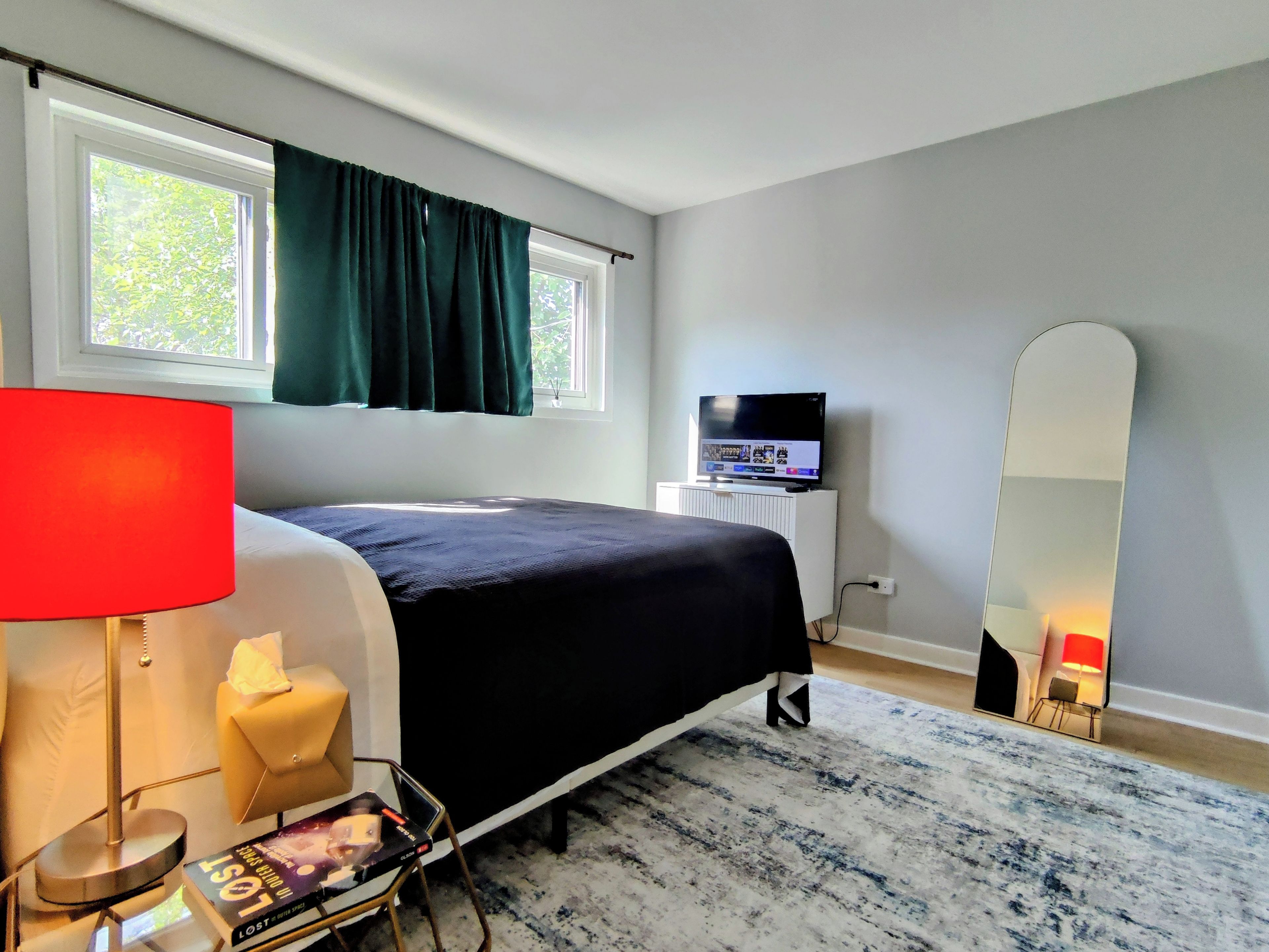 A hotel room with a bed, a television, a bedside table with a lamp, a chair, and a large mirror.