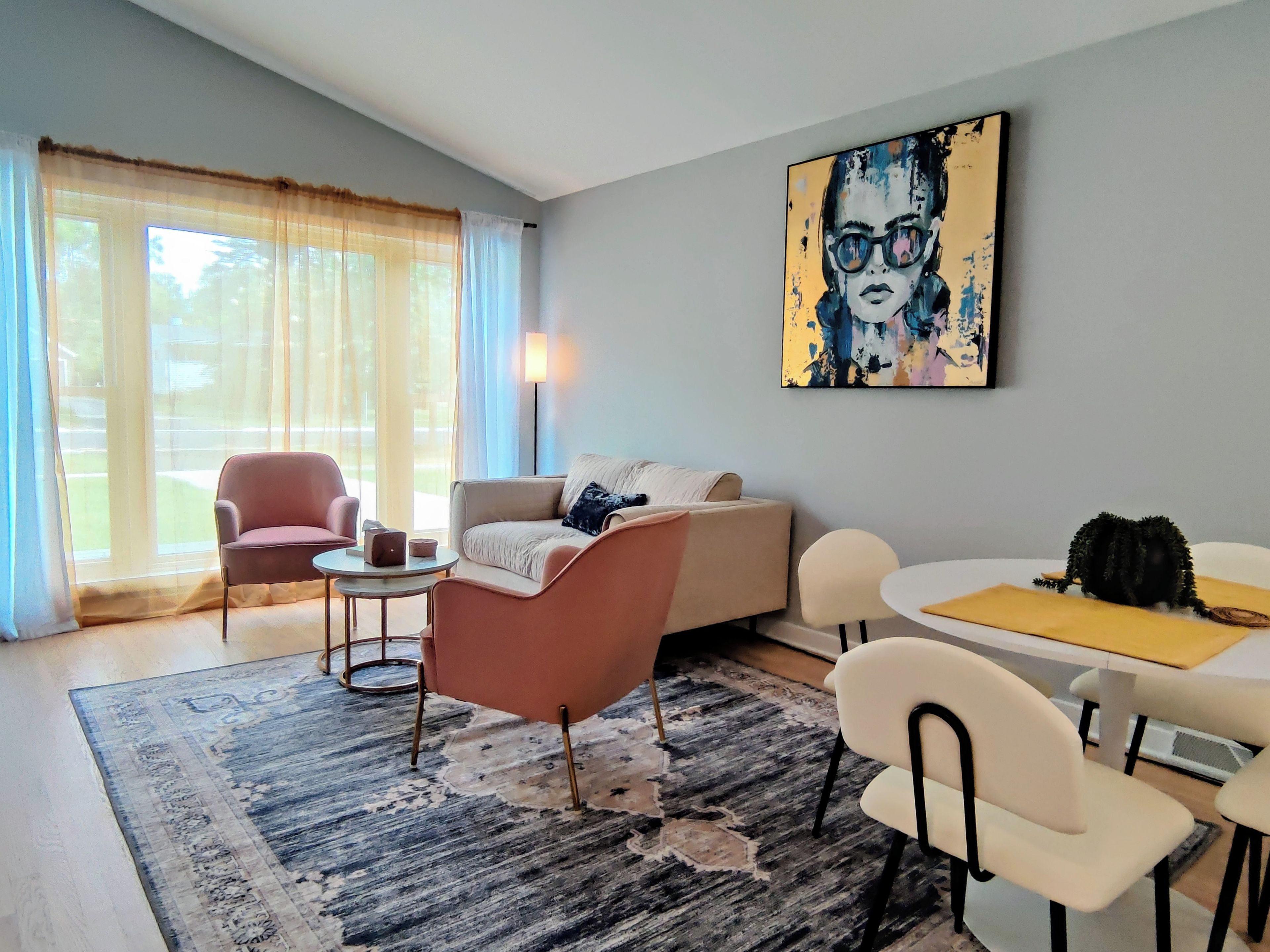 A modern living room with a sofa, armchairs, a coffee table, and a dining table. There is a large window with curtains, a painting on the wall, and a rug on the floor.