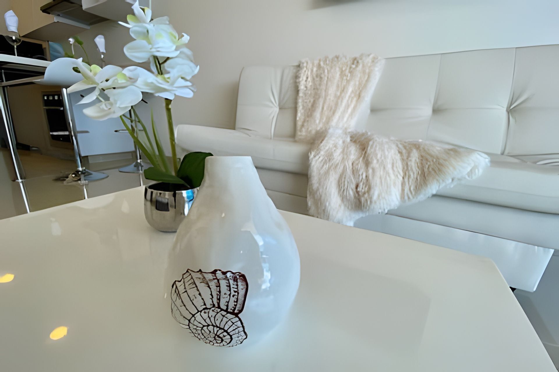 A white vase with a shell design on a white table, a white sofa with a fluffy throw, and white orchids in a metal pot.