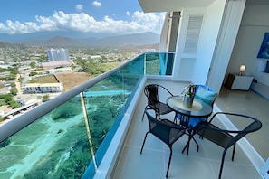A balcony with a glass railing overlooking a coastal landscape, a table with chairs, and a view of distant buildings. - Apartamentos Reserva del Mar - by SOHO (Santa Marta)