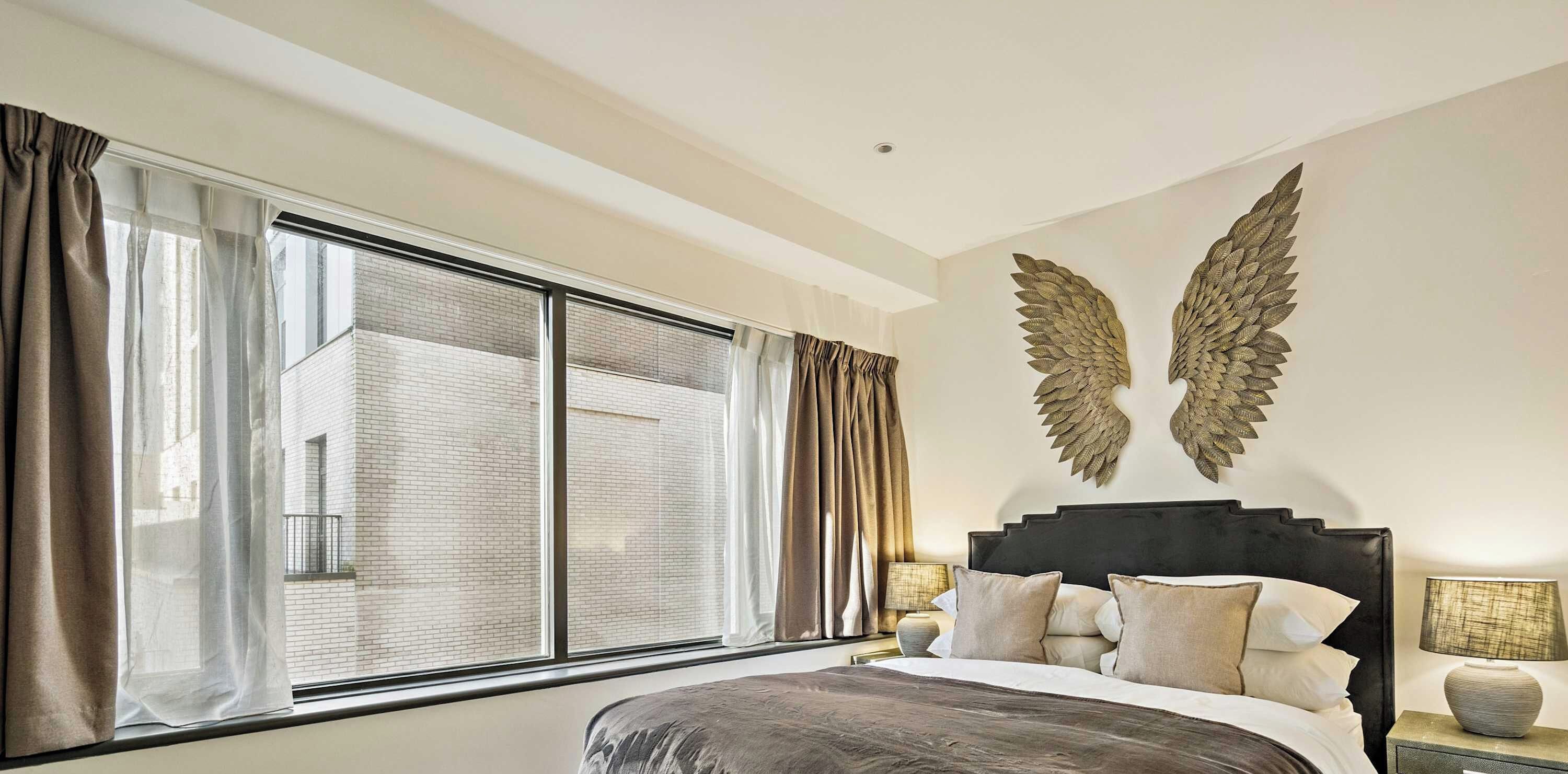 A hotel room with a large window, a bed with a dark headboard, and two wall-mounted decorative wings.