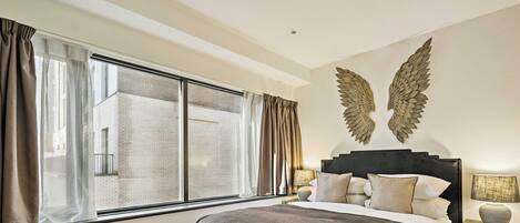 A hotel room with a large window, a bed with a dark headboard, and two wall-mounted decorative wings.