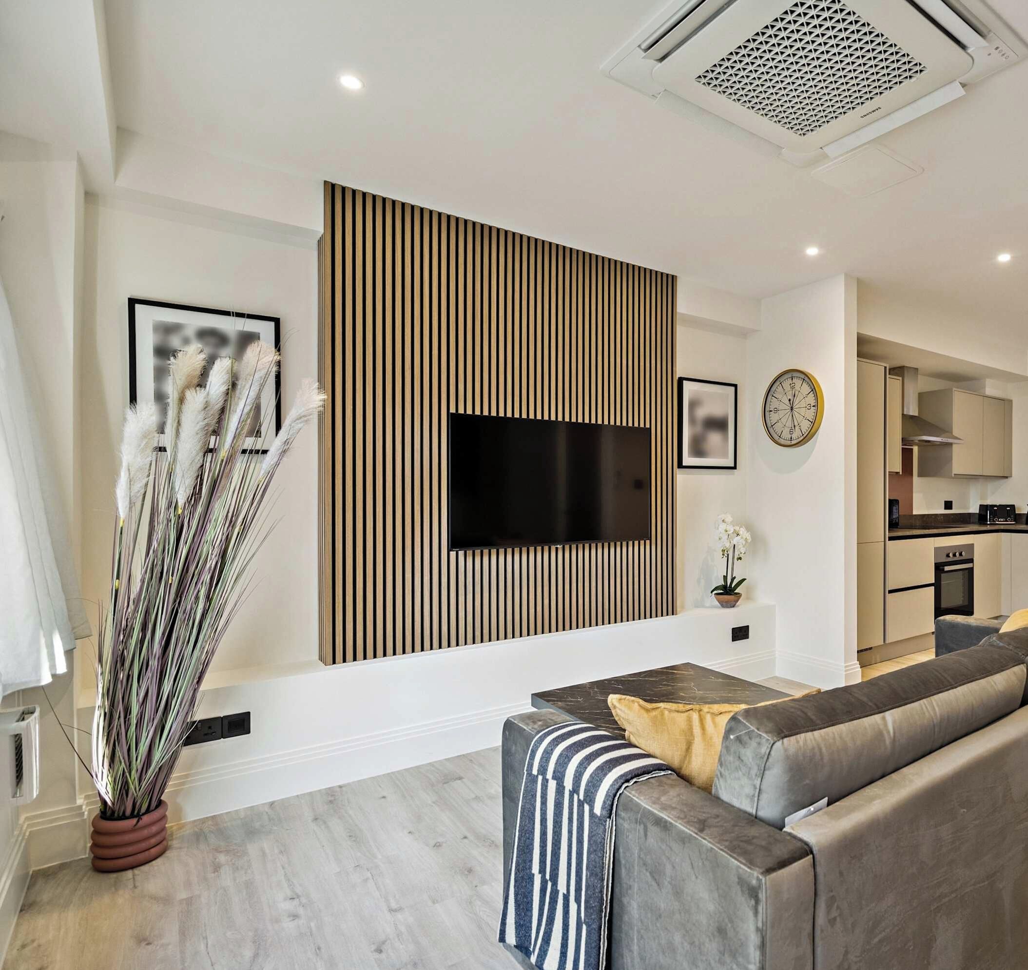 A modern living room with a flat-screen TV mounted on a wooden panel wall, a grey sofa, a coffee table, and a potted plant.