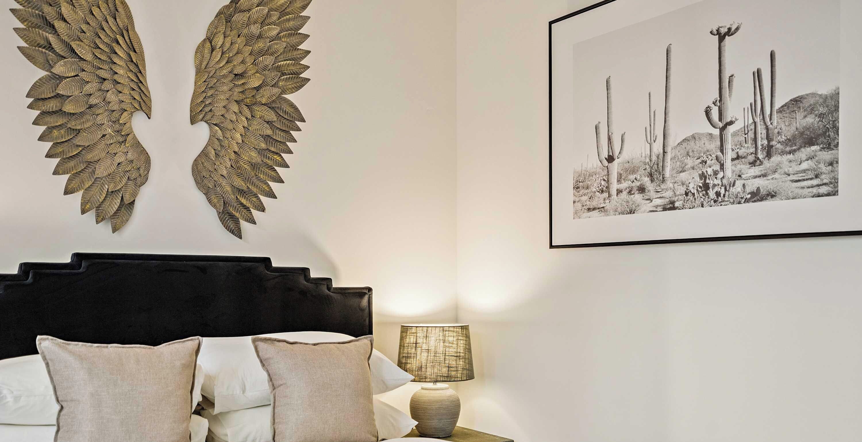 A bedroom with a bed, pillows, a wall-mounted wing-like decoration, and a framed picture of cacti.