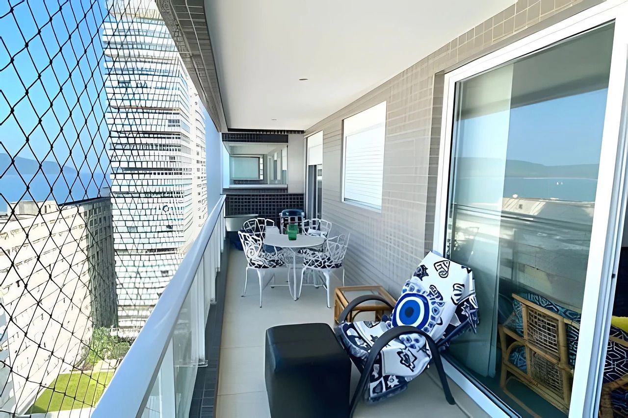 A balcony with a dining area, a sofa, and a large window offering a view of a building and the sea.