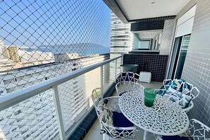 A balcony with a white metal railing, a round table with chairs, and a view of the cityscape through a chain-link fence. - SHB - A Beira-Mar no Gonzaga (Santos)