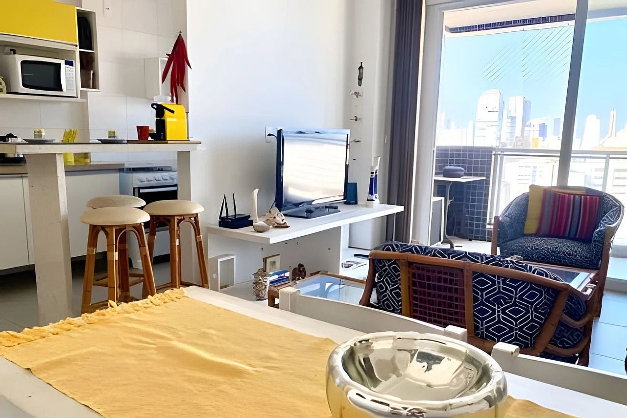 A modern living space with a kitchen area, a dining table with stools, a large window with a city view, and a comfortable seating area with patterned cushions.