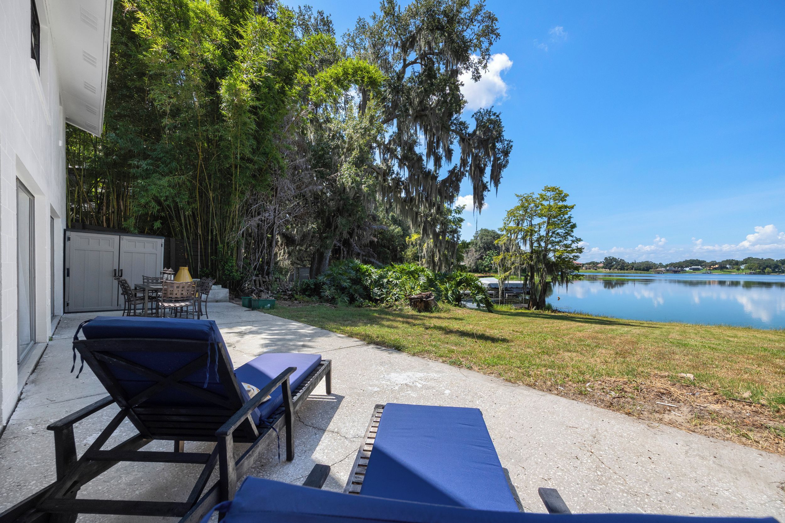 Un patio con sillones de descanso y una mesa, con vista a un lago con árboles y un edificio al fondo.