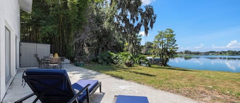 Un patio con sillones de descanso y una mesa, con vista a un lago con árboles y un edificio al fondo.