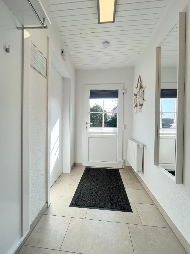 A hallway with a black rug, white walls, a white door with a window, and a ceiling light.