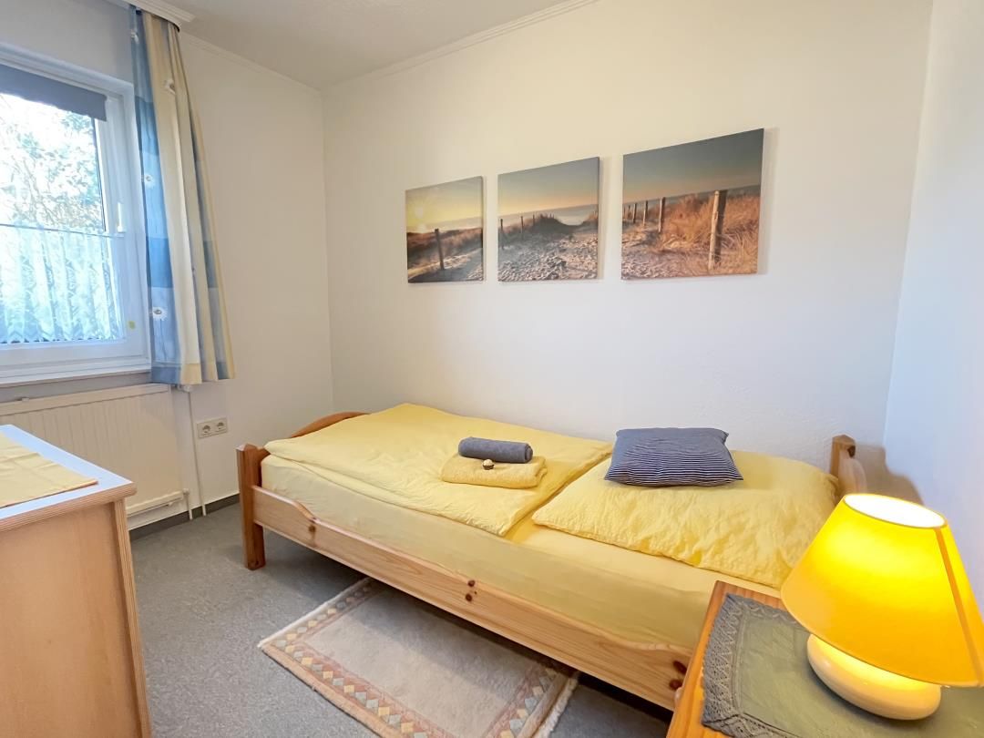 Un lit simple avec une couvre-lit jaune, une tête de lit en bois et une table de chevet avec une lampe. Trois tableaux sont accrochés au mur et représentent des scènes de plein air.
