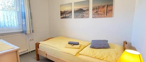 A single bed with a yellow bedspread, a wooden headboard, and a bedside table with a lamp. There are three wall-mounted pictures depicting outdoor scenes.