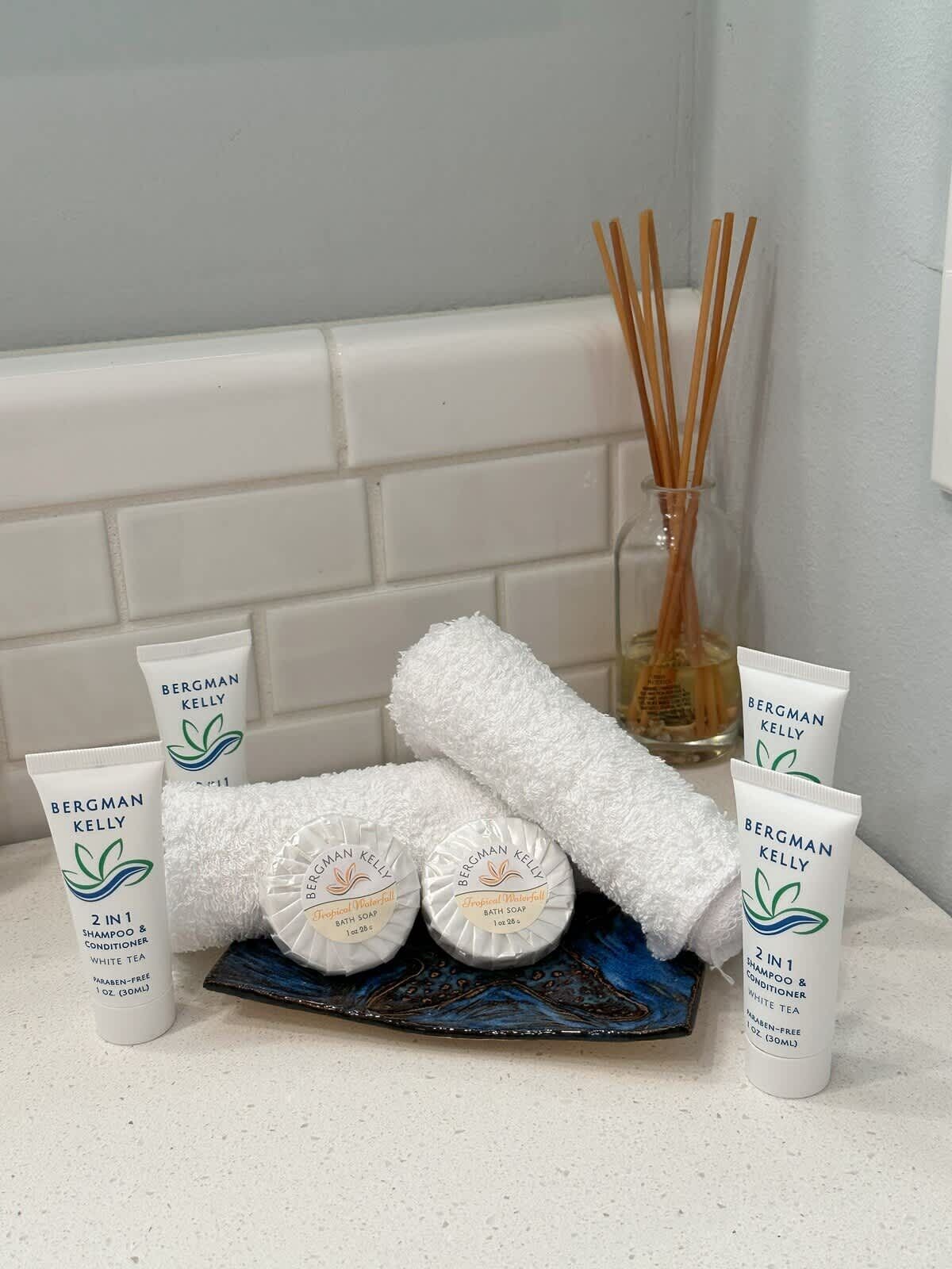 A bathroom with toiletries including Bergman Kelly shampoo, conditioner, and bath soap, a white towel, and a glass bottle with reed diffusers.