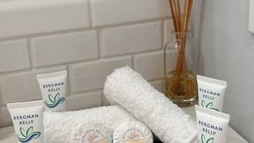 A bathroom with toiletries including Bergman Kelly shampoo, conditioner, and bath soap, a white towel, and a glass bottle with reed diffusers.