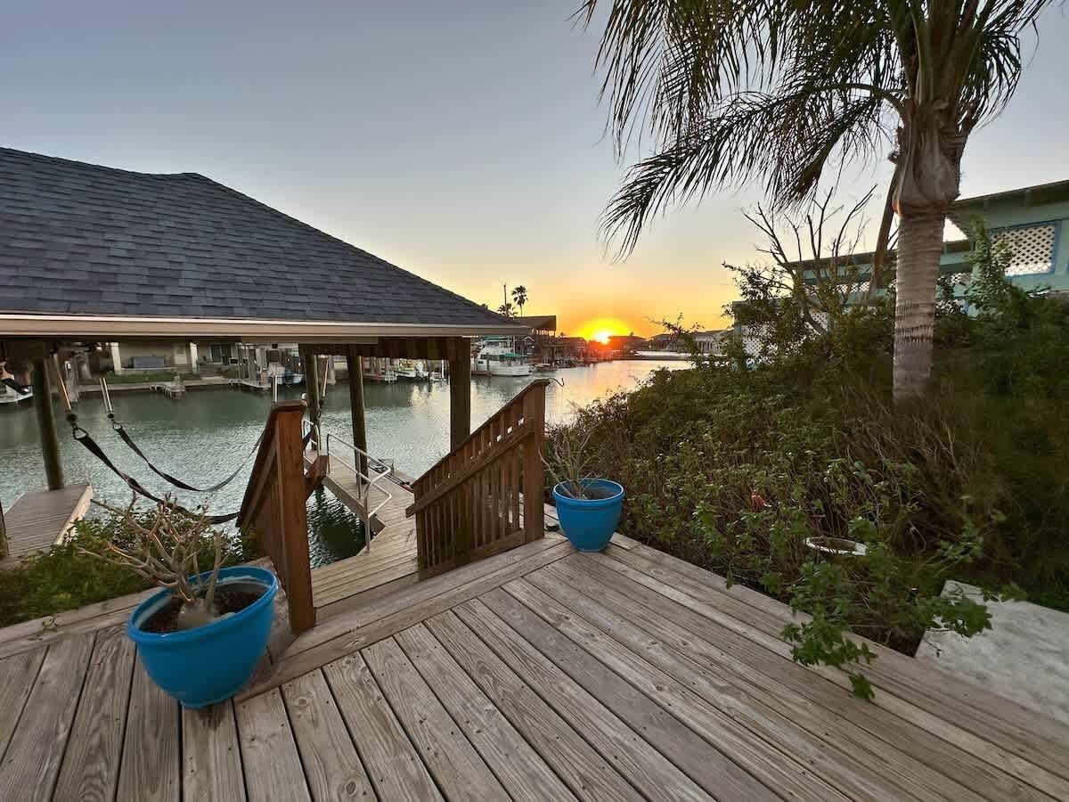 Une terrasse en bois offrant une vue sur un coucher de soleil sur l’eau, un espace abrité et deux plantes en pot.