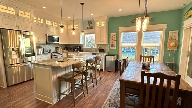 A modern kitchen with stainless steel appliances, a central island with bar stools, and a dining area with a wooden table and chairs. - Isla Bella - Canal Retreat w Private Dock & Water Views (Port Isabel)