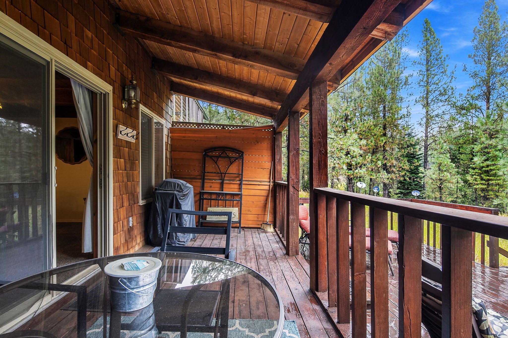 Une terrasse en bois avec une table et des chaises en verre, un barbecue couvert et une vue sur les arbres.