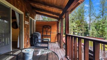 Une terrasse en bois avec une table et des chaises en verre, un barbecue couvert et une vue sur les arbres.