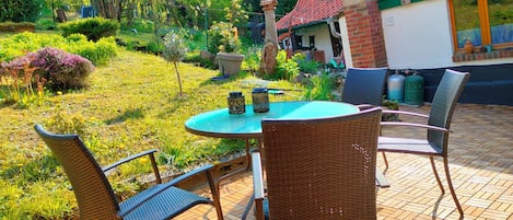 An outdoor patio with a round table and wicker chairs, surrounded by a garden with various plants and a red-tiled building in the background.