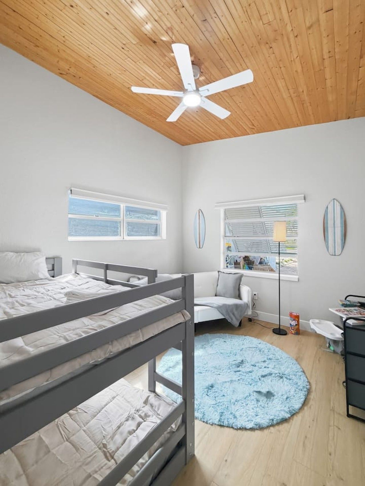 A bedroom with a bunk bed, a ceiling fan, a round blue rug, and a window with a view.