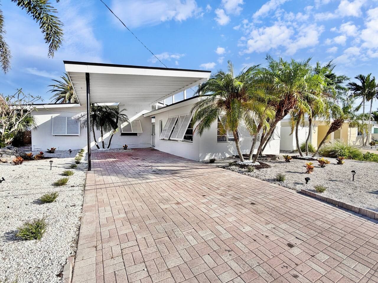 A modern house with a covered patio, a paved driveway, and a garden with palm trees.