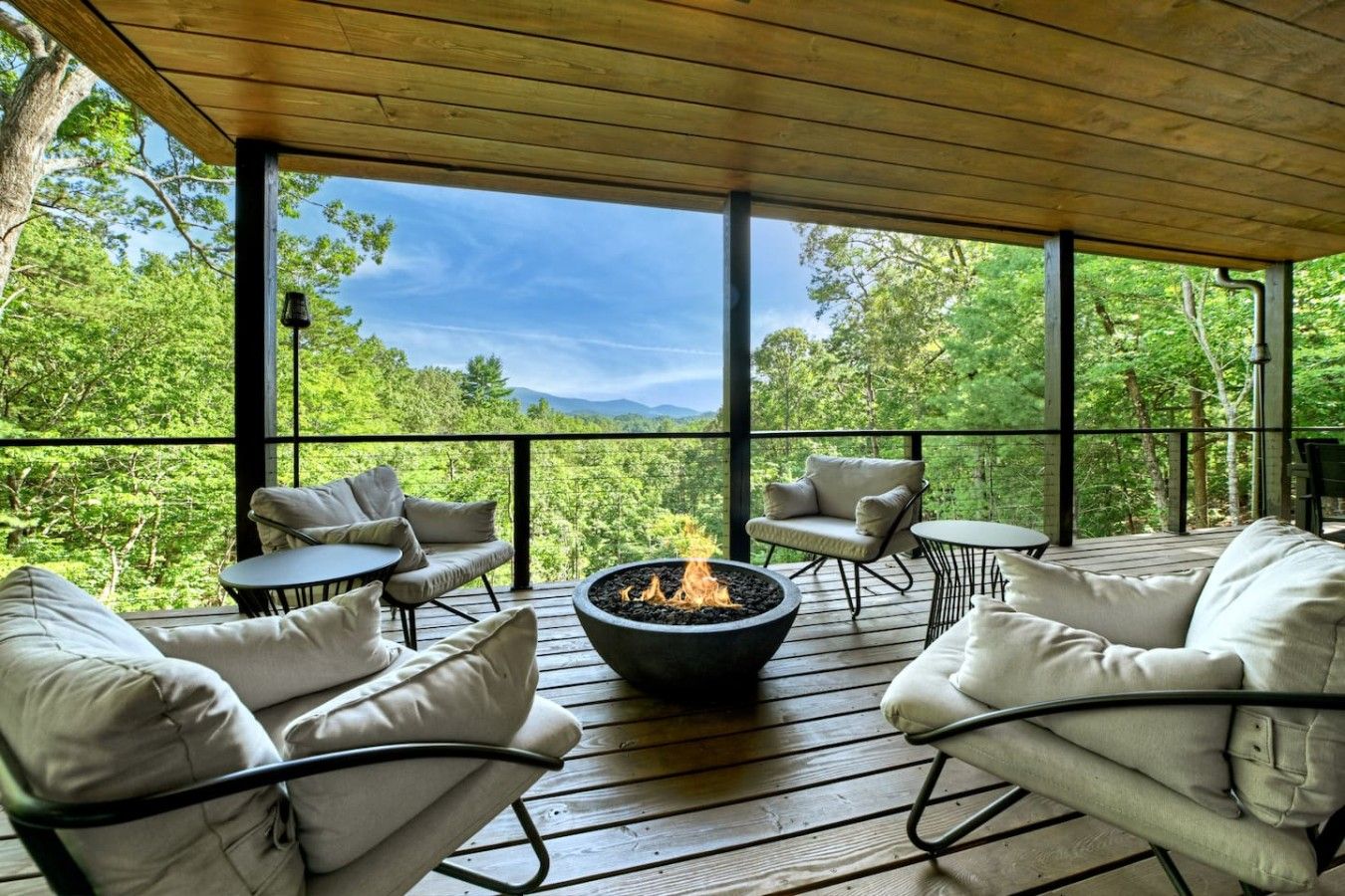 A covered outdoor seating area with a fire pit, surrounded by cushioned chairs and tables, overlooking a forested landscape.