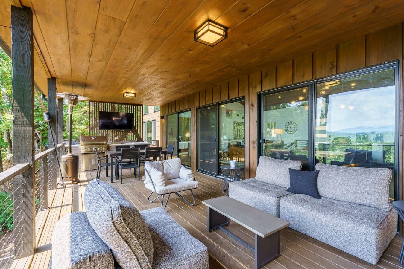 Une terrasse en bois, dotée de mobilier d’extérieur, d’un barbecue et de grandes fenêtres offrant une vue dégagée.