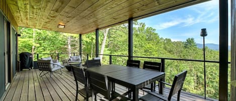 A covered outdoor deck with a table and chairs, overlooking a forested area.