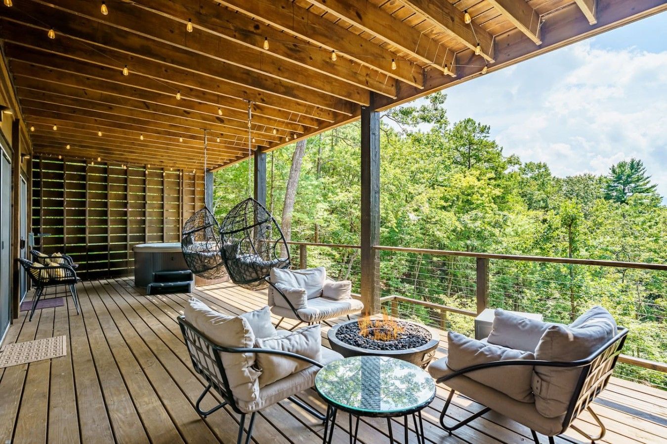 A covered outdoor deck with seating, a fire pit, and a view of a forest.