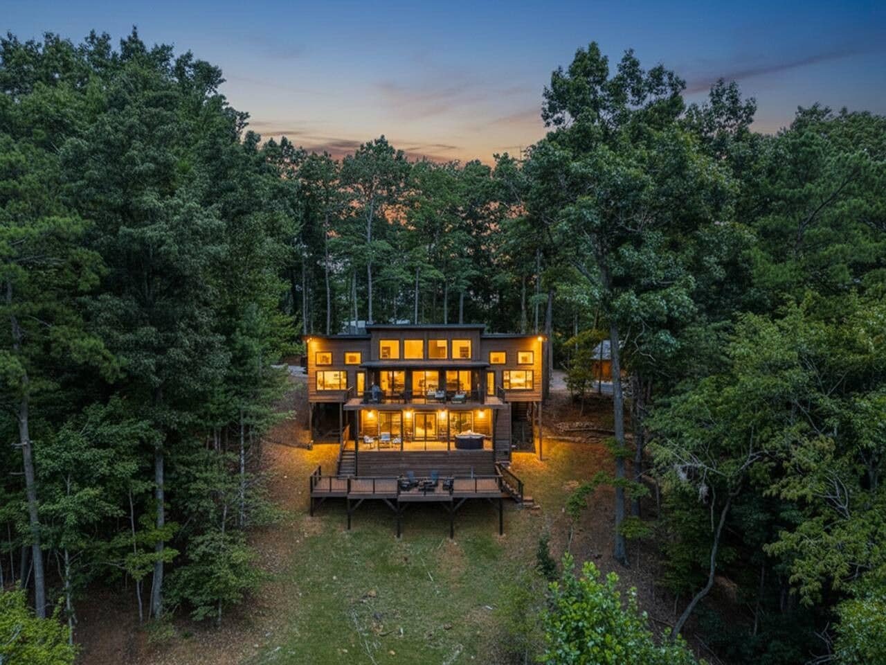 A modern two-story house surrounded by dense forest, with a deck and outdoor seating area.