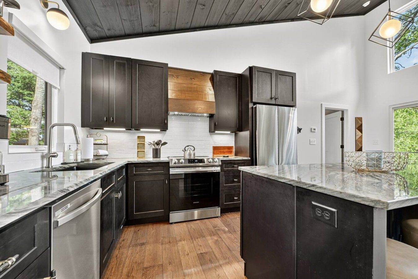 A modern kitchen with dark cabinetry, stainless steel appliances, and a wooden floor.