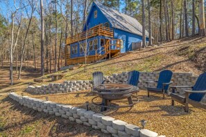 A blue cabin with a deck, surrounded by a stone fire pit and Adirondack chairs.