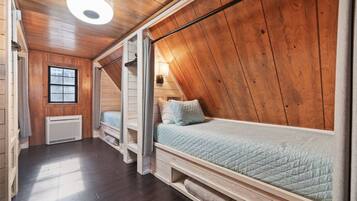 Une pièce avec un plafond en pente, des murs en bois, un lit avec un matelas matelassé, une fenêtre et un luminaire fixé au mur.
