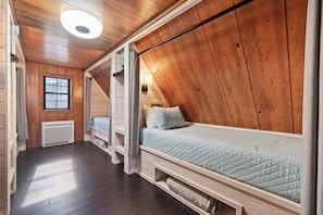 A room with a sloped ceiling, wooden walls, a bed with a quilted mattress, a window, and a wall-mounted light fixture.