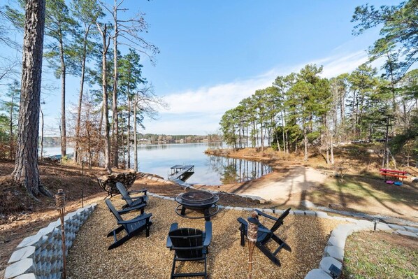 A lakeside outdoor seating area with Adirondack chairs, a fire pit, and a dock extending into the water.