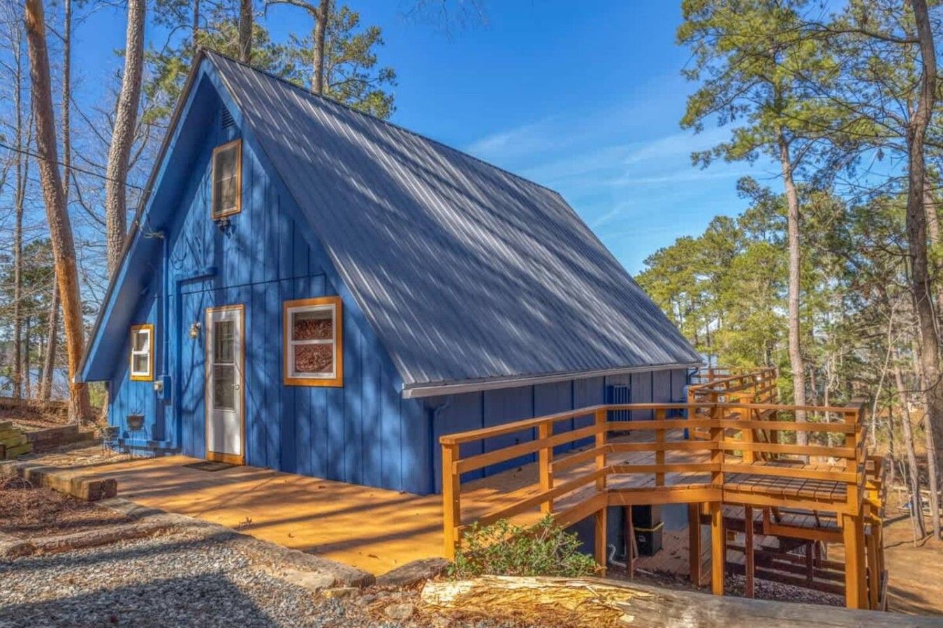 A blue cabin with a wooden deck and a metal roof, surrounded by trees.