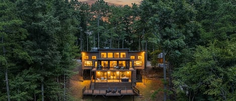 A modern two-story house surrounded by dense forest, with a deck and outdoor seating area.