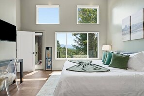 A bedroom with a large bed, a TV, and a view of trees and mountains through the windows. - Mountain Haus | 4BR | 3 Decks | Views | Fireplaces (Blue Ridge)