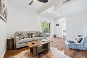 A spacious living room with a grey sofa, a blue armchair, a wooden coffee table, and a large window offering a view of greenery outside. - Mountain Haus | 4BR | 3 Decks | Views | Fireplaces (Blue Ridge)