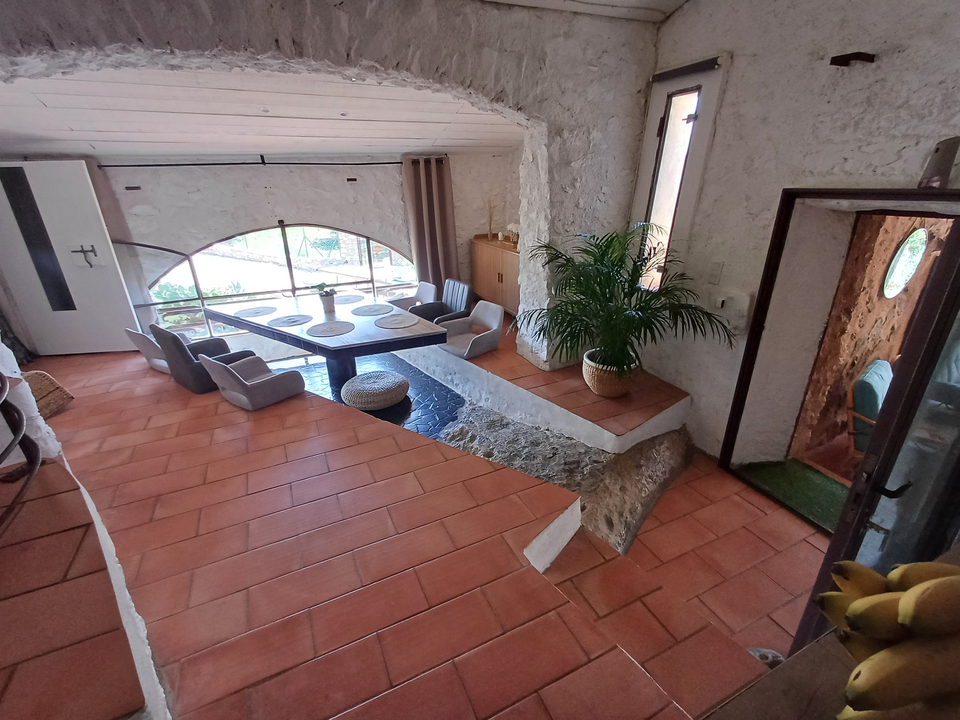 A room with a dining area, seating, a potted plant, and a view through a window.