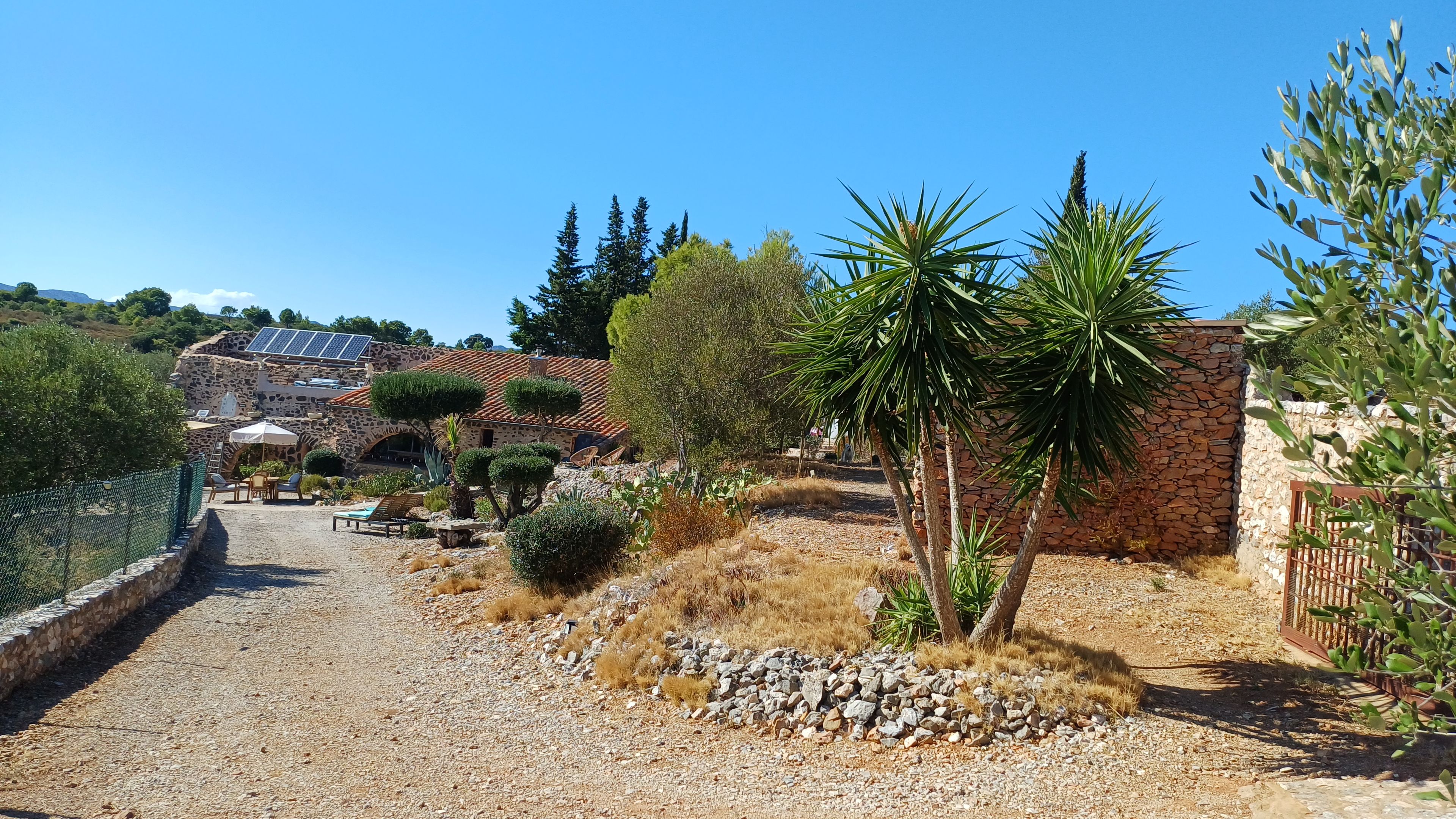 A rustic stone building with a tiled roof, surrounded by a gravel path, a palm tree, and various shrubs.