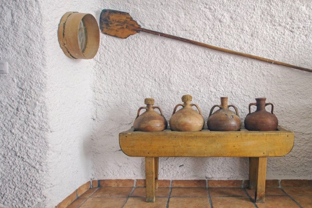 A rustic interior with a wooden shelf holding four pottery items and a wooden spoon hanging on the wall.