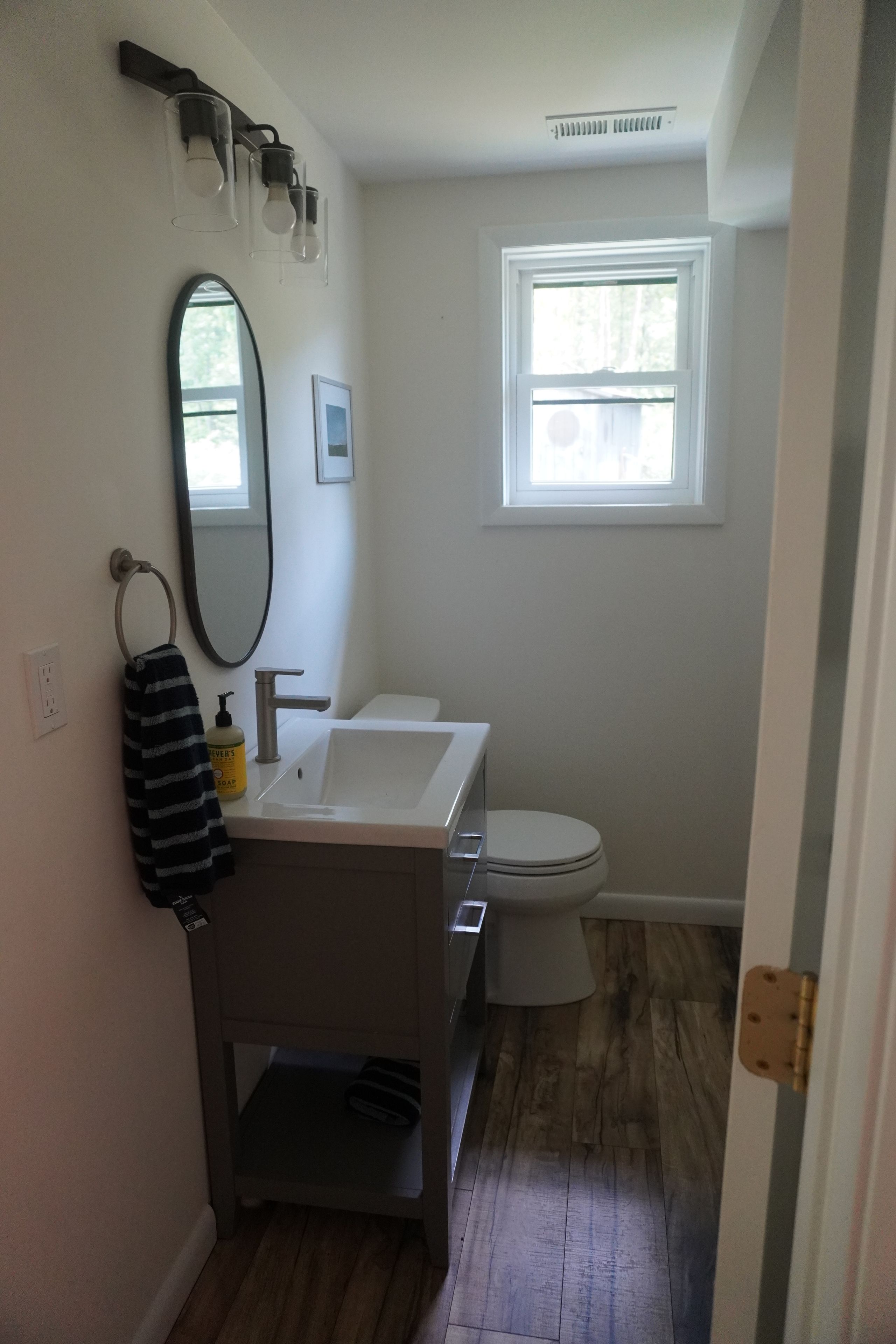 A bathroom with a white sink, mirror, and toilet. There is a window with blinds, a towel rack, and a soap dispenser on the sink.