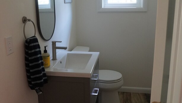 Ein Badezimmer mit weißem Waschbecken, Spiegel und Toilette. Ein Fenster mit Jalousien, ein Handtuchhalter und ein Seifenspender befinden sich am Waschbecken.