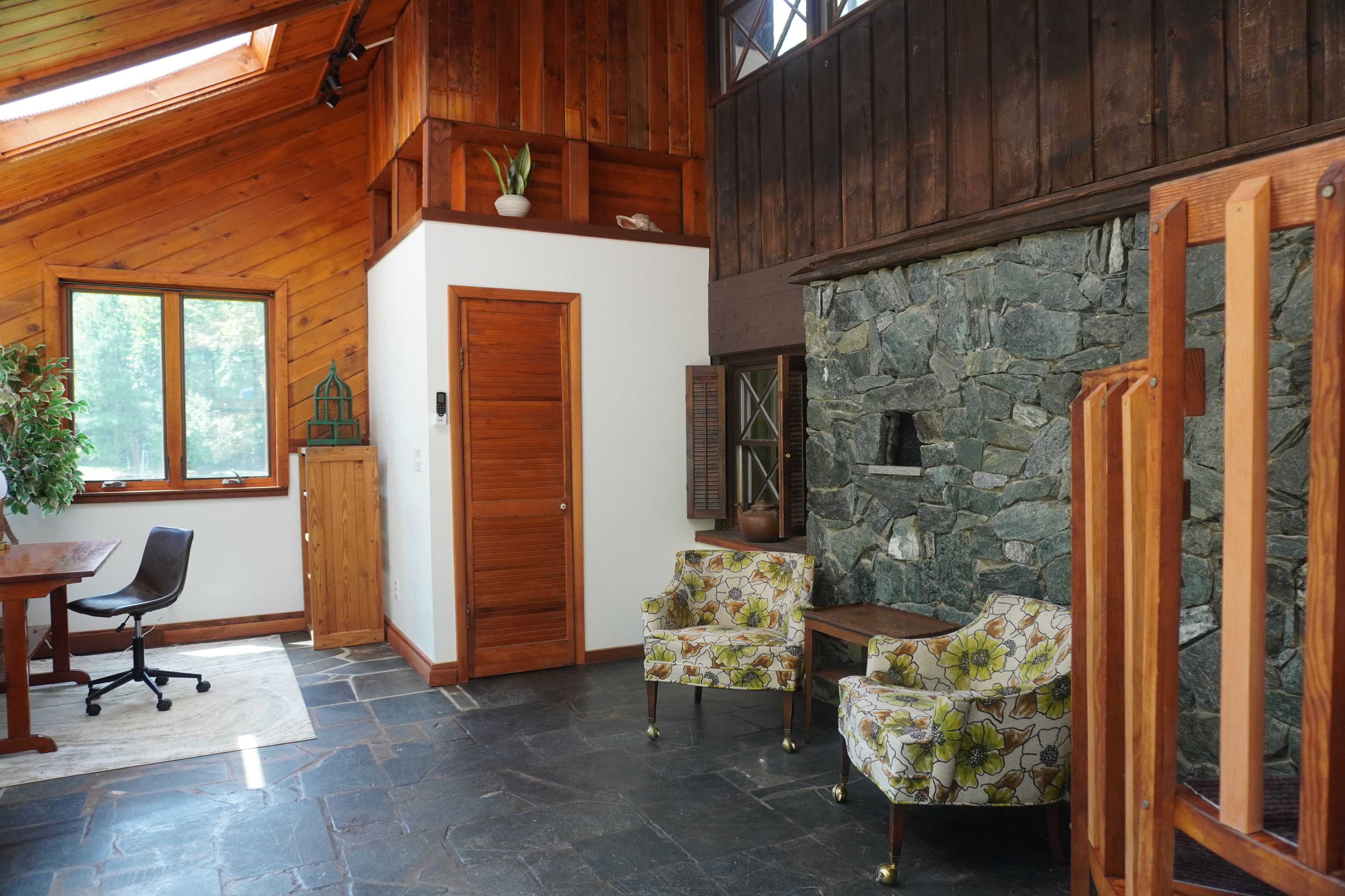 A room with a stone wall, wooden ceiling, and a door. There is a chair and a table with a plant.