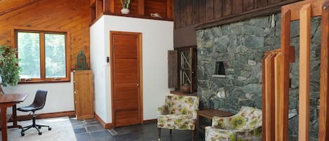 A room with a stone wall, wooden ceiling, and a door. There is a chair and a table with a plant.
