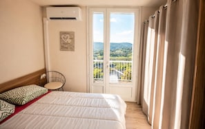 Quarto com cama, poltrona, aparelho de ar condicionado e varanda com vista para vegetação e colinas.