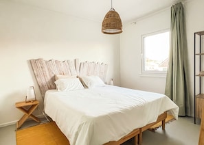 Un dormitorio con estructura de cama de madera, ropa de cama blanca, una lámpara colgante tejida, una mesita de noche y una ventana con cortinas.
